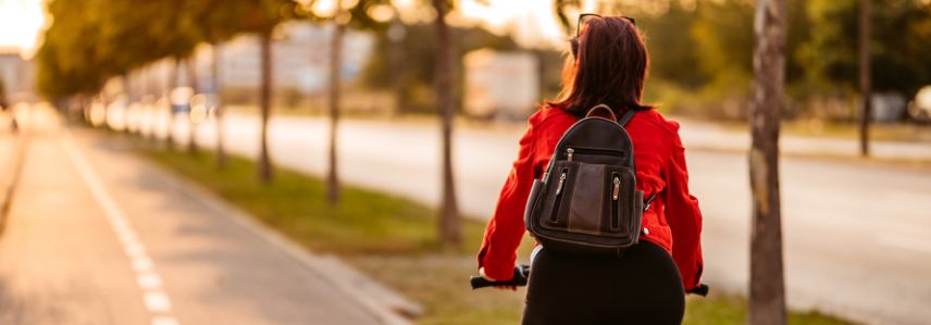 Person cycling on a city bike path during sunset, wearing a red jacket and backpack, with trees lining the street.